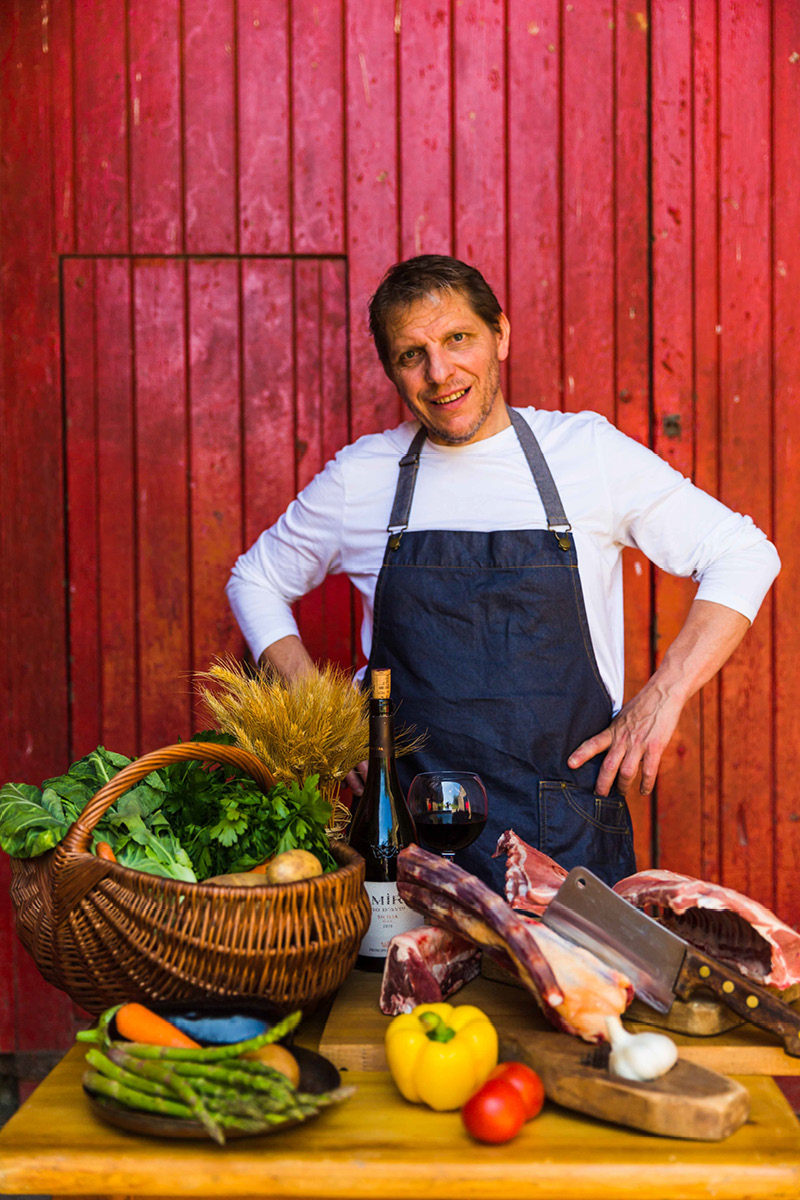 Chef Patron at La Locanda, Maurizio Bocchi, stood behind a table with fresh meat and vegetables, a bottle of Italian wine and full glass.